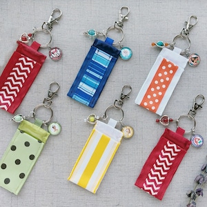 May include: A collection of keychains with fabric pouches in various colors and patterns. Designs include red with white zigzags, blue stripes, white with orange polka dots, green with black polka dots, yellow stripes, and red with white zigzags.