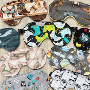 May include: A collection of eight fabric sleep masks featuring various patterns including cowboy boots and hats, dinosaurs, video game controllers, camouflage, comic book text, baseballs, space astronauts, science equipment, and stormtrooper helmets.