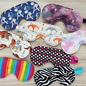 May include: A collection of seven sleep masks featuring various patterns including a blue Eiffel Tower print, a purple galaxy design, a gray fox motif, a unicorn print, a horse pattern, a black and pink polka dot design, a rainbow stripe, and a zebra print.