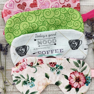 May include: Four fabric sleep masks with elastic bands. The masks feature various patterns, including hearts, green swirls, floral designs, and a coffee-themed print with the text "Today's good MOOD is sponsored by COFFEE."