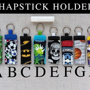 May include: Eight fabric chapstick holders with metal keyrings, labeled A through H. Patterns include green pixels, soccer balls, bat symbols, a dinosaur, a skull, a fighter pilot theme, a basketball, and a viking. A white lip balm is centered above.