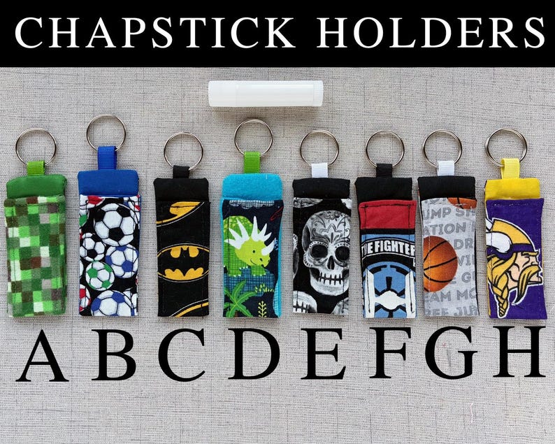 May include: Eight fabric chapstick holders with metal keyrings, labeled A through H. Patterns include green pixels, soccer balls, bat symbols, a dinosaur, a skull, a fighter pilot theme, a basketball, and a viking. A white lip balm is centered above.