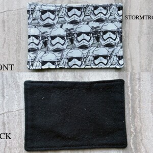 May include: A rectangular fabric patch featuring a repeating black and white pattern of Stormtrooper helmets. The front side displays the graphic print with the text STORMTROOPER, while the back side is a solid, plain black fabric material.