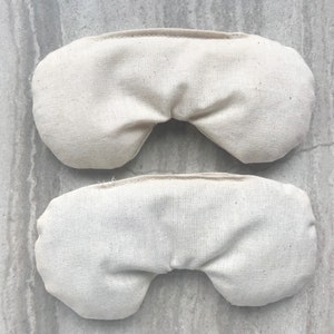 May include: Two beige fabric eye pillows, one on top of the other. The pillows are rectangular and have a curved shape in the middle. They are made of a soft, natural fabric.