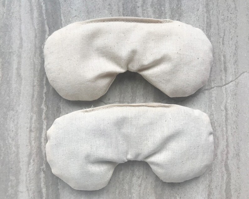 May include: Two beige fabric eye pillows, one on top of the other. The pillows are rectangular and have a curved shape in the middle. They are made of a soft, natural fabric.