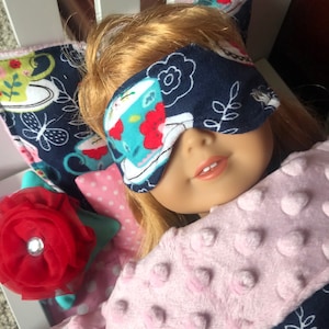 May include: A doll with light brown hair wears a dark blue sleep mask featuring a colorful tea cup and floral pattern. The doll is tucked under a soft, light pink textured blanket with raised dots, resting against a pillow with a large red fabric flower.