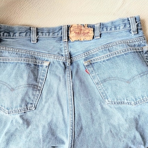 May include: Light blue denim jeans with classic five-pocket styling. Features include a red tab on the back pocket and a worn leather patch. The jeans have a faded appearance and are likely vintage.