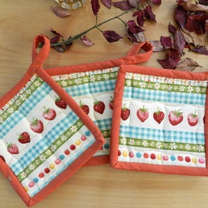 Peut inclure: Trois maniques matelassées rouge et blanc avec un motif de fraises. Les maniques ont une poignée en boucle et sont bordées d'orange.
