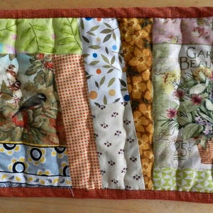 May include: Rectangular quilted placemat with various fabric patterns. Features floral, bird, and dotted designs in green, orange, and pink. A solid rust-coloured border surrounds the piece, with the text "GARDEN BEAUTY" visible.