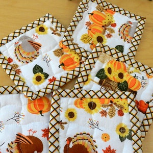 May include: Set of six quilted fabric pot holders with a Thanksgiving theme. The pot holders feature a white background with images of turkeys, pumpkins, sunflowers, and autumn leaves. The borders are a brown and yellow checkered pattern.