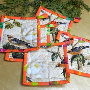 May include: Set of four quilted pot holders featuring a bird and floral design. The pot holders have a bright orange border and are made from a cream-coloured fabric.