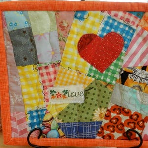 May include: A colourful patchwork quilt featuring a red heart applique and the word "love" embroidered on a small patch. The quilt is made from a variety of patterned fabrics, including gingham and floral designs, and is framed with an orange border. The quilt is a square shape.