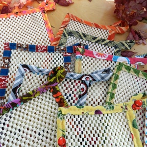 May include: A set of nine colourful fabric-trimmed pot holders with a white mesh centre. The pot holders are made of different fabrics with various patterns and colours, including red, blue, green, yellow, and white.