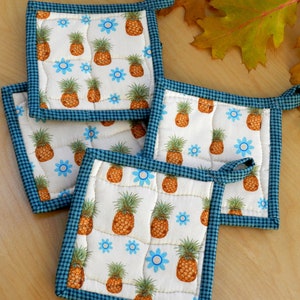May include: Four quilted pot holders with a blue and white checked border. The pot holders feature a white background with a repeating pattern of pineapples and blue flowers.