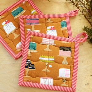 May include: Three quilted pot holders with a pink and white striped border. The pot holders are made of a brown fabric with a pattern of colourful cakes on stands.