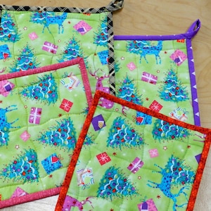 May include: Three green quilted pot holders with a Christmas tree and reindeer design. The pot holders have different coloured borders: pink, purple, and black and white plaid.