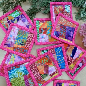 May include: A set of ten pink and white fabric coasters with a patchwork design. The coasters are made of different fabrics with various patterns and colours.