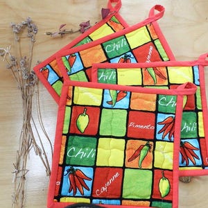 Peut inclure: Ensemble de trois maniques matelassées rouge et orange avec un motif de piment. Les maniques ont un motif à carreaux avec les mots "Chili", "Pimento", "Cayenne" et "Hili" imprimés dessus.