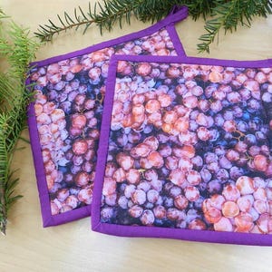 Peut inclure: Deux maniques carrées avec un motif de raisin. Le tissu présente un gros plan de raisins violets et roses. Chaque manique a une bordure violette et une boucle pour suspendre. Les maniques mesurent environ 20 cm de côté.