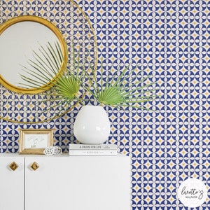 Puede incluir: Una pared blanca con un papel tapiz de patrón geométrico azul y amarillo. El papel tapiz presenta un patrón repetitivo de círculos y diamantes. Un espejo dorado, un jarrón blanco con plantas verdes y una estantería blanca con dos libros y un marco de fotos están frente a la pared.