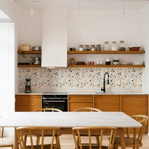 Puede incluir: Una cocina moderna con gabinetes blancos, una campana extractora blanca y un salpicadero de azulejos de terrazo. La cocina cuenta con una encimera de madera, un fregadero de acero inoxidable y un grifo negro. Hay dos estantes de madera sobre la encimera, llenos de frascos y platos. Una mesa de comedor de mármol blanco con cuatro sillas de madera se encuentra en el centro de la habitación.