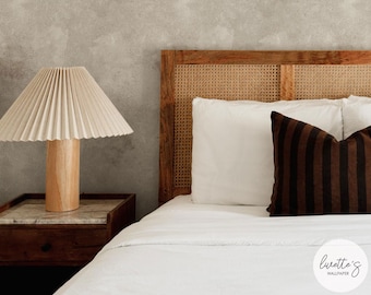 Può includere: Una scena di camera da letto con una testiera in legno con dettagli in canna intrecciata, biancheria da letto bianca e un cuscino a righe marroni. Un paralume plissettato beige è posto su una base di lampada in legno su un comodino con piano in marmo. Il muro ha una finitura grigia strutturata.