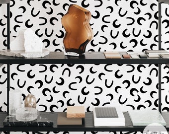 Puede incluir: Una estantería de metal negro exhibe varios objetos decorativos sobre un fondo blanco con un patrón curvo negro repetido. Los artículos incluyen un jarrón de vidrio marrón, esculturas de piedra blanca y pequeños bloques de madera.