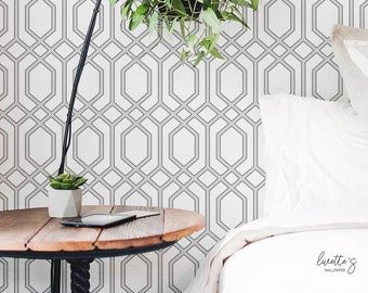 May include: A bedroom scene featuring a geometric patterned wallpaper in white and gray. A hanging plant with green leaves is suspended in front of the wall. A wooden side table with a black lamp and a small succulent plant is in the foreground.