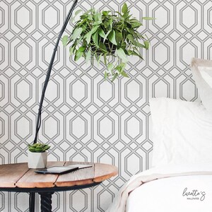 May include: A bedroom scene featuring a geometric patterned wallpaper in white and gray. A hanging plant with green leaves is suspended in front of the wall. A wooden side table with a black lamp and a small succulent plant is in the foreground.