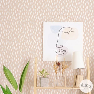 May include: A beige wallpaper with a white polka dot pattern. A framed abstract line drawing of a face with pastel colours is hanging on the wall. A wooden shelf with a potted plant, dried flowers in a glass vase, and a white lamp is in front of the wall.