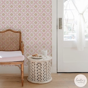 Op de afbeelding: Roze en goudkleurig behang met patroon op een muur, met een stoel en een kleine, ronde tafel. De tafel heeft een witte beker en een bord met eten. Een witte deur en gordijn zijn op de achtergrond. Behang van Lunette's Wallpaper.
