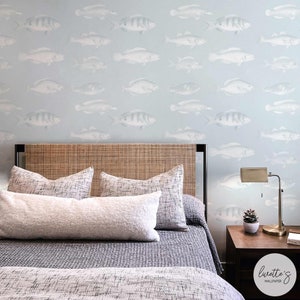 Könnte beinhalten: Ein Schlafzimmer mit hellblauer Wand, die mit einem sich wiederholenden Fischmuster verziert ist. Das Bett hat ein gewebtes Kopfteil, gemusterte Kissen und eine graue Tagesdecke. Eine Messinglampe und eine kleine Pflanze stehen auf einem Nachttisch aus Holz.