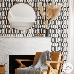 May include: A modern living room with a round gold mirror, a brown leather armchair, and a gray blanket. The wall features a black and white chain-link pattern wallpaper. A black vase with dried pampas grass sits on the mantel.