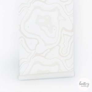 May include: A roll of white wallpaper with a light gray, marbled pattern. The wallpaper is partially unrolled, revealing the design. The bottom edge is rolled up. The brand name "Lurette's Wallpaper" is in the bottom right corner.