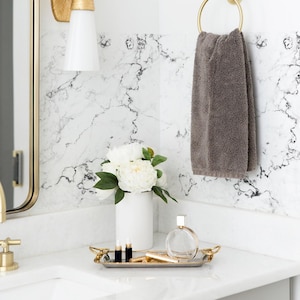 May include: A white bathroom vanity with a gold faucet, a white ceramic vase with white flowers, a gold tray with a clear glass bottle and gold tubes, and a gray towel hanging on a gold ring.