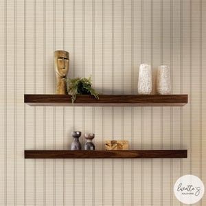 Puede incluir: Dos estantes flotantes de madera marrón oscuro montados en una pared cubierta con papel tapiz beige con finas rayas verticales. El estante superior sostiene una escultura de rostro abstracto dorado y una planta verde. El estante inferior sostiene dos jarrones pequeños y una caja de madera.