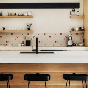 Puede incluir: Una cocina moderna con encimera blanca, fregadero negro y grifo negro. El protector contra salpicaduras presenta un estampado floral. Estantes de madera con platos y plantas. Taburetes negros en primer plano. El diseño es minimalista y elegante.