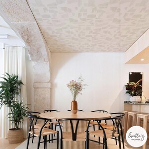 Könnte beinhalten: Ein Esszimmer mit einem runden Holztisch und schwarzen Stühlen mit gewebten Sitzen. Die Decke ist mit hellem geometrischem Tapetenmuster verziert. Eine hohe Pflanze in einem geflochtenen Korb und eine Vase mit Trockenblumen ergänzen das Dekor.