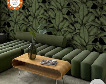 Op de afbeelding: Een moderne woonkamer met een houten salontafel, een groene bank en een vaas met een plant. De achtergrond is voorzien van behang met een groot groen bladerpatroon. De kamer heeft een eigentijds design.