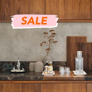 May include: A kitchen scene with wooden cabinets and countertops. The word "SALE" is displayed in orange on a pink background. Decorative items include vases, bottles, glasses, and cutting boards. A small fountain sculpture is on a black plate.