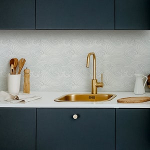 Op de afbeelding: Een gouden keukenkraan met een enkele hendel en een witte porseleinen gootsteen met een witte golfpatroon achterwand.