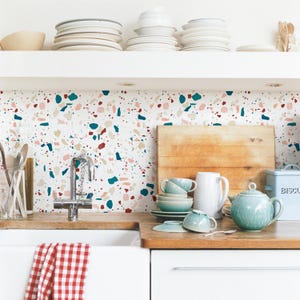 Può includere: Un rivestimento da cucina con un motivo terrazzo in tonalità rosa, rosso, blu e beige. Il rivestimento è dietro un lavello da cucina bianco con un rubinetto cromato. Ci sono piatti e una teiera sul bancone.