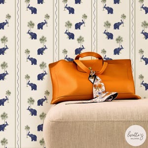 Pode incluir: Uma mala de couro laranja com um lenço estampado repousa sobre uma otomana bege. O fundo apresenta um papel de parede com um padrão repetido de elefantes azuis e palmeiras verdes sobre um fundo creme.