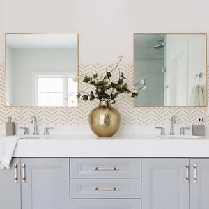May include: A white bathroom vanity with two sinks, gold taps, and grey cabinets. The vanity has a white countertop and gold hardware. The backsplash is white with a gold geometric pattern. A gold vase with green plants sits on the countertop.