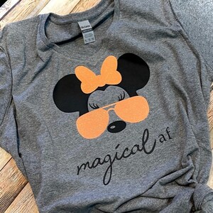 May include: Gray t-shirt with a black and orange cartoon image of Minnie Mouse wearing sunglasses and a bow. The text "magical af" is printed below the image.