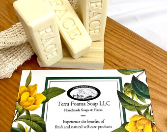 May include: Three white bars of soap with the word "FACE" embossed on each bar. The soap bars are arranged on a wooden stand. The stand is on a wooden surface. A white card with green trim and a floral design is in the foreground. The card has the text "Terra Foama Soap LLC" and "Handmade Soaps & Fizzes".