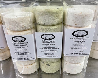 May include: Five clear plastic containers with white shower steamers inside. The containers are labeled with the names of the scents: Orange & Rosemary, Herbs de Provence, Energy Shower Steamers, Energy Shower Steamers, and Energy Shower Steamers. Each container has a label with the name of the scent, the number of steamers, and the essential oil blend.