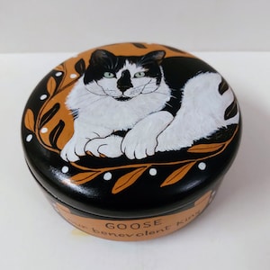 May include: A round, black painted box with a hand-painted image of a black and white cat. The cat is lying down with green eyes. The box has a gold-colored border with leaves and the words "GOOSE benevolent king".