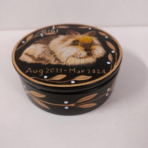 May include: A round, black decorative box with a hand-painted image of a fluffy rabbit on the lid. The lid also features the name "Gigi" and the dates "Aug 2011-Mar 2024". Gold-colored leaves and white dots decorate the box.