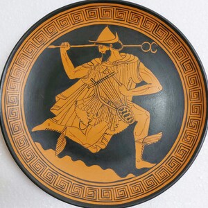 May include: A round ceramic plate featuring a black and orange Greek-style illustration of a bearded figure wearing a pointed hat and winged boots. The figure holds a long staff and carries a lyre, surrounded by a geometric meander border pattern.